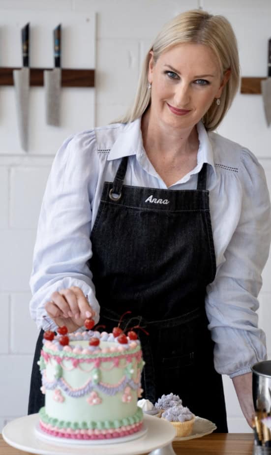 A lady with blonde hair and blue eyes called Anna puts the finishing touches on a vintage style cake with cherries