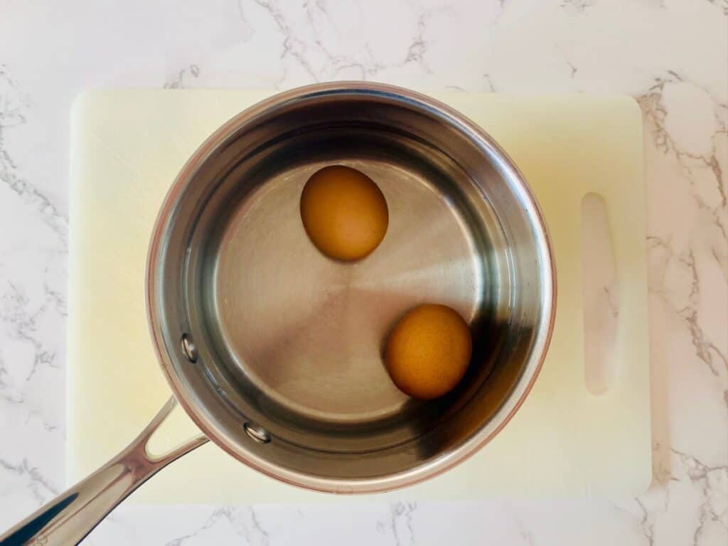 A saucepan with 2 eggs and water on a bench