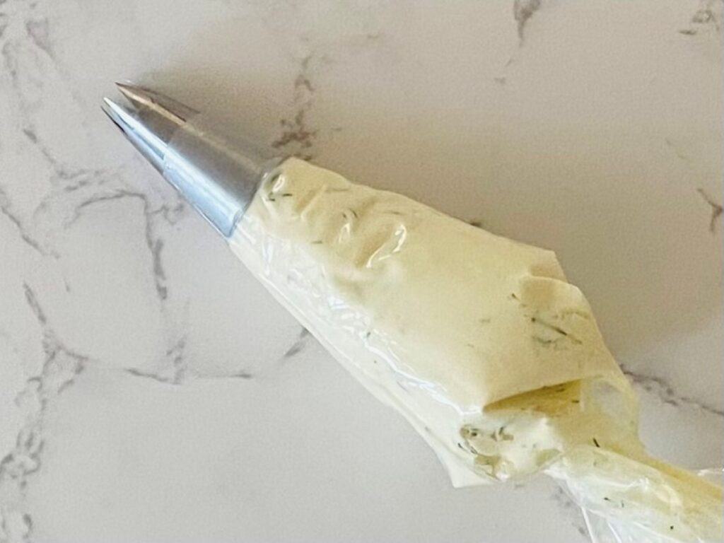 A piping bag filled with dill and horseradish cream cheese sits on a marble bench ready for Smoked Salmon Pikelets.