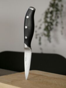 A stainless steel kitchen knife with a black handle standing on a wooden cutting board in a kitchen setting.