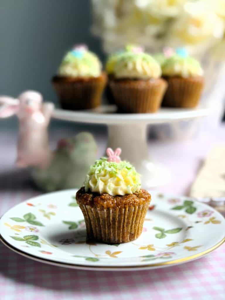 A single Easter-themed mini carrot cake on a Wedgwood wild strawberry plate with more cupcakes behind on a cake stand